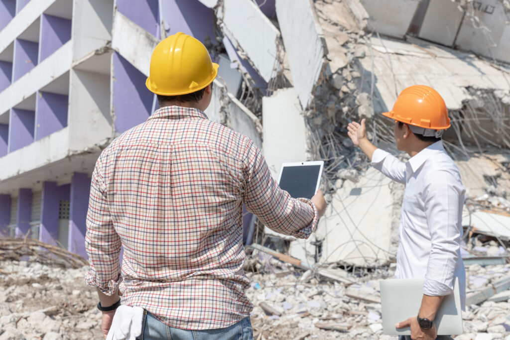 Two workers in hard hats outside a partially collapsed building.