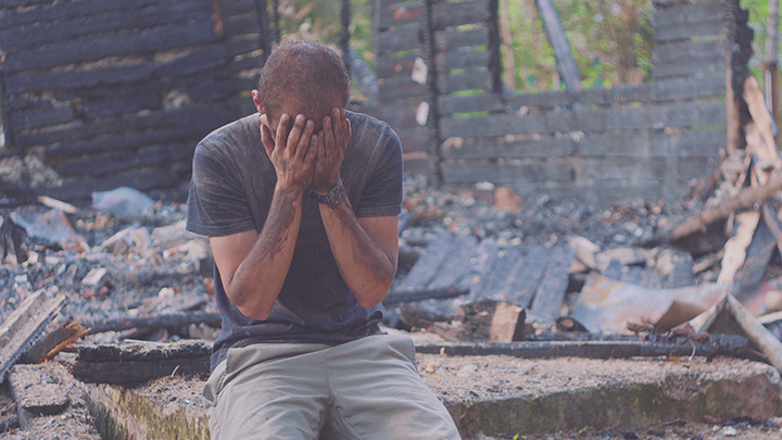 Photo of a man with his hands on his face, sitting in front of a fire damaged building.