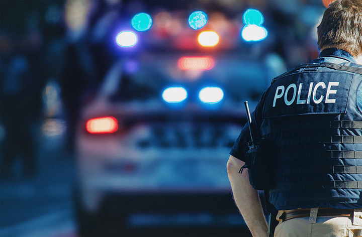 Photo of the back of a police officer in front of their patrol car with lights on.