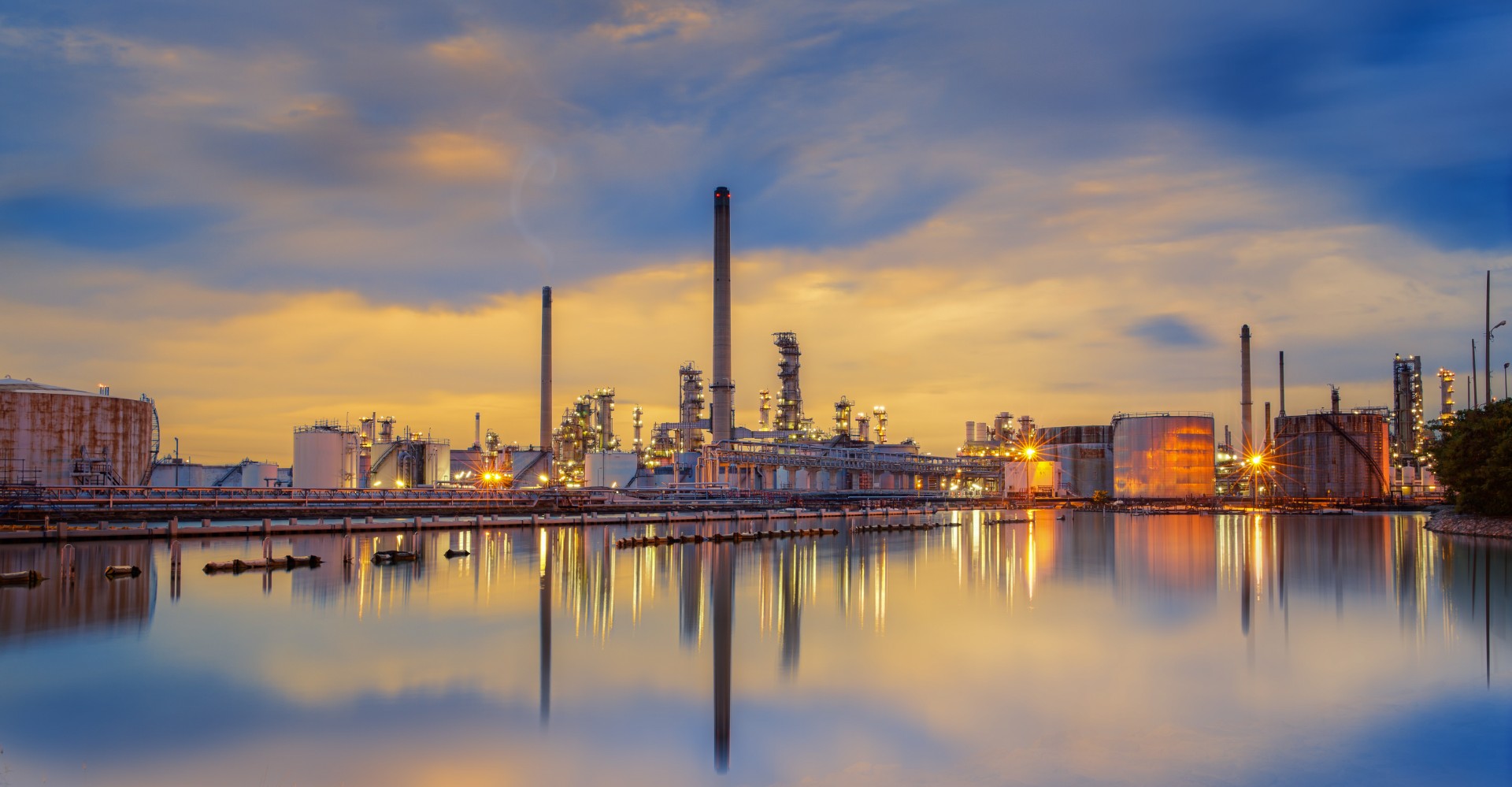 An oil refinery at dusk, its lights and smokestacks reflected in the still water in front of it.