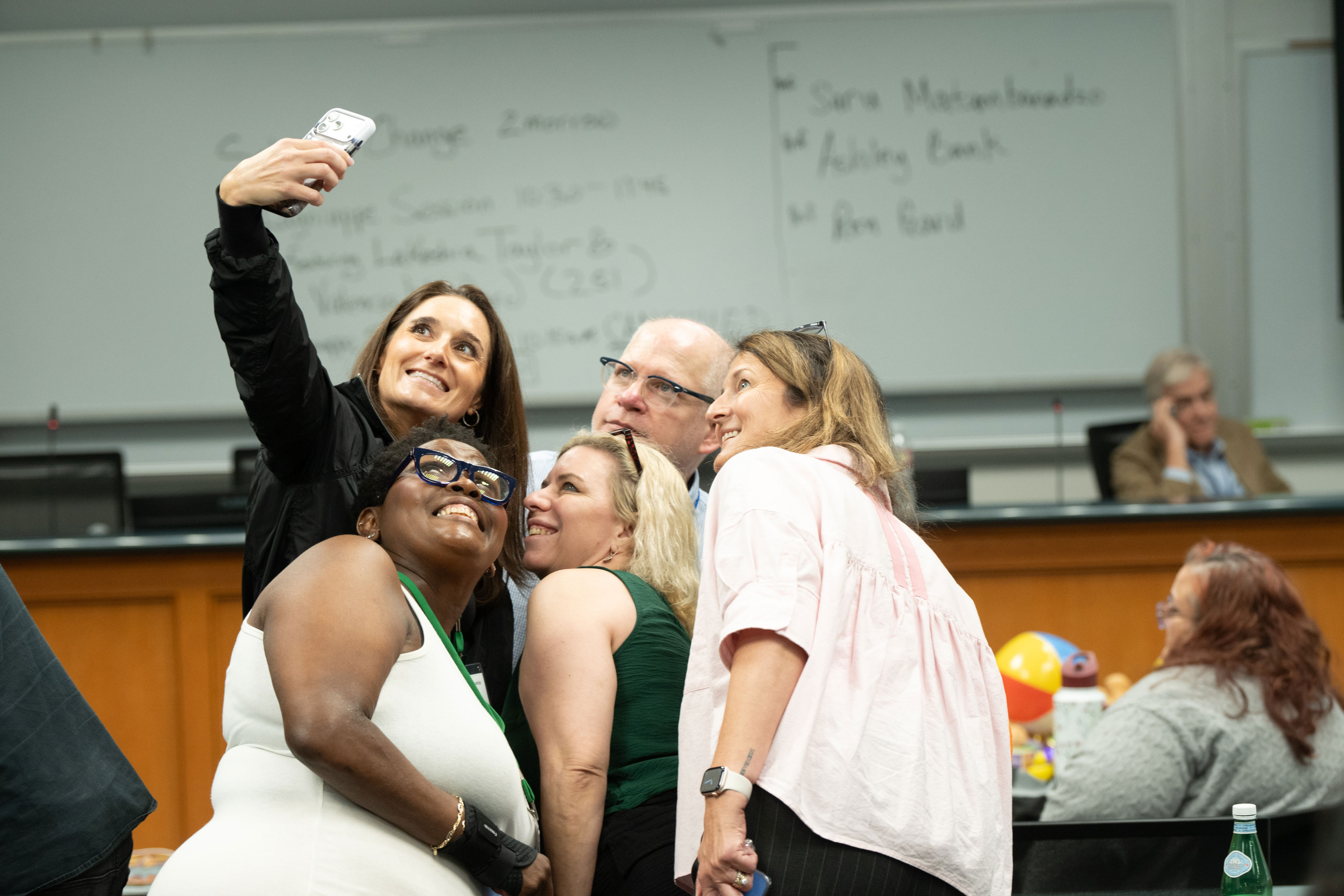 group of six people pose together for a selfie taken by a woman holding up her phone. They're smiling and looking up at the camera in a Tulane Law lecutre hall