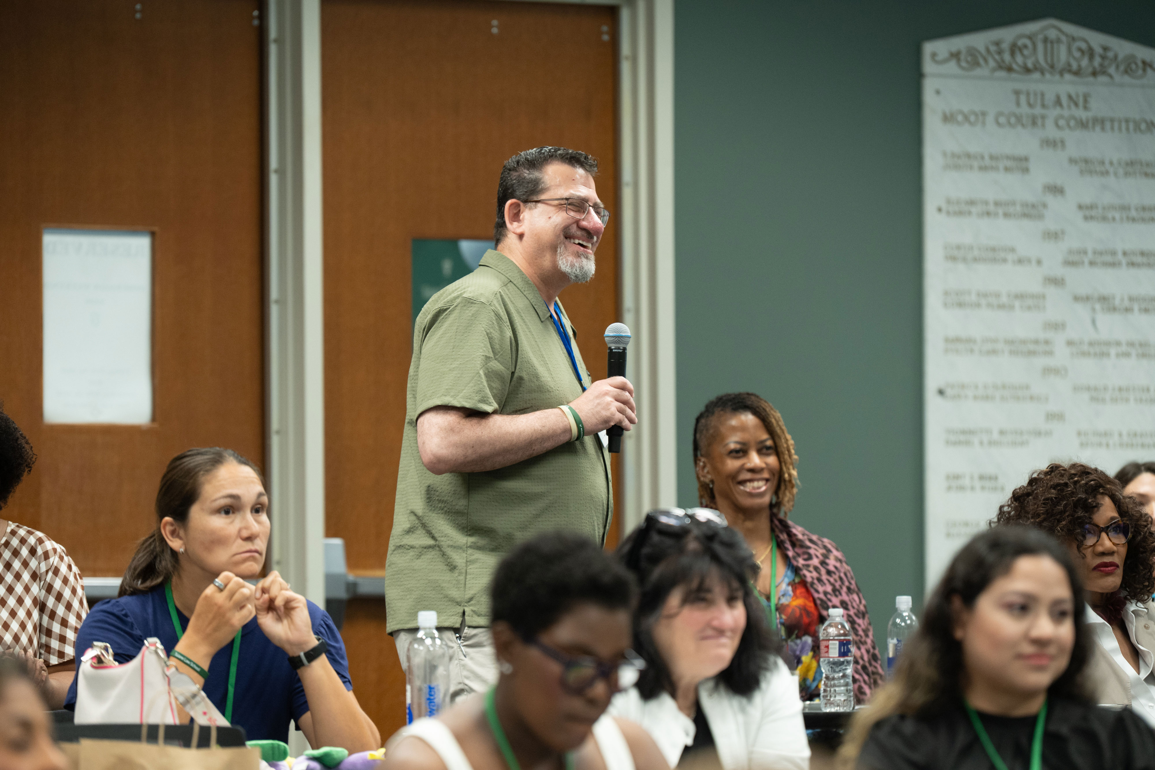 : A man with glasses and a gray beard, wearing an olive shirt, stands and speaks into a handheld microphone in a Tulane Law Classroom