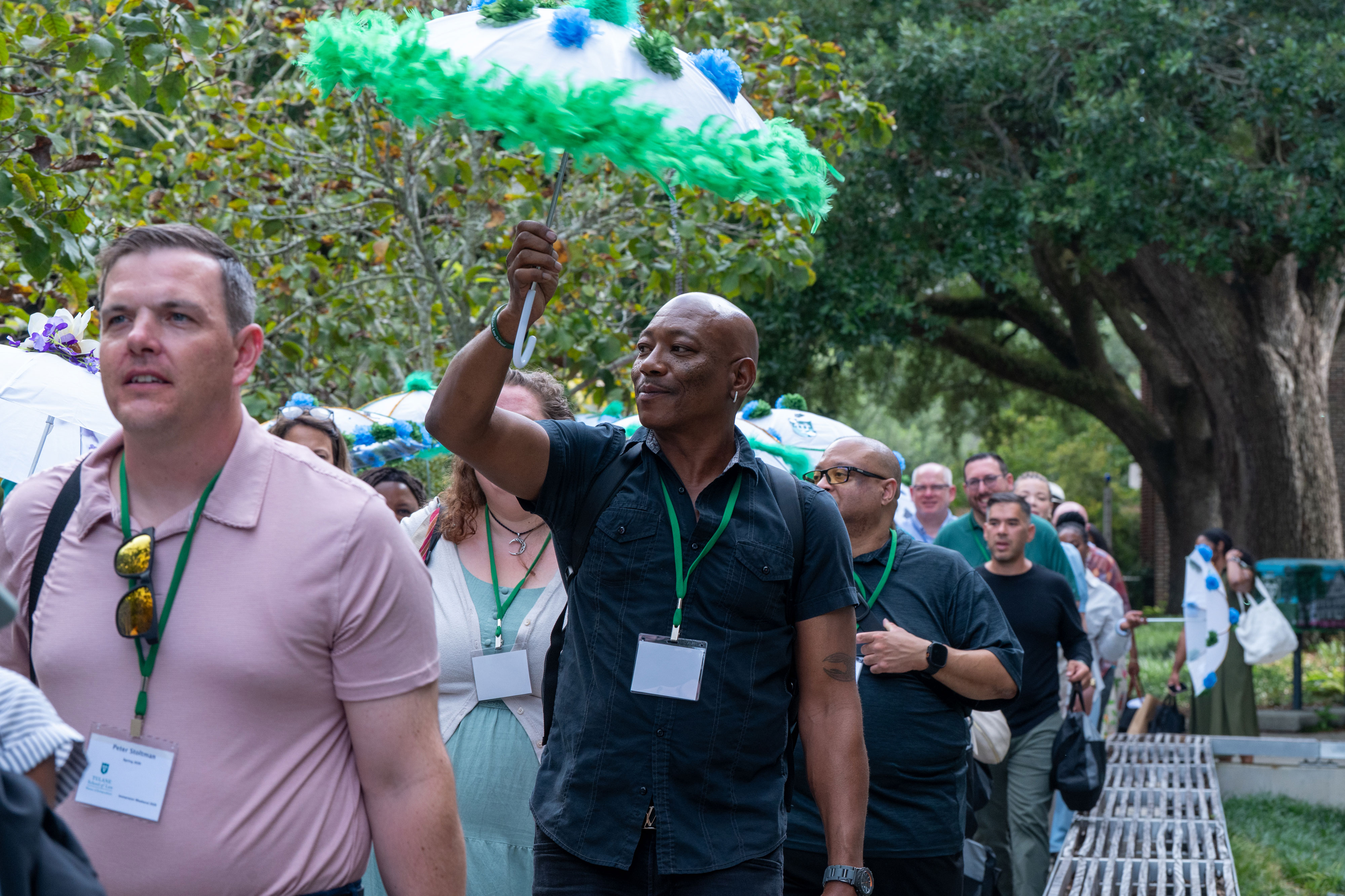 A group of people walking on Tulane uptown campus, lanyards around their necks. In the foreground, a man in a dark shirt holds up a decorative umbrella covered in green and blue feathers, smiling as he looks off to the side. Others behind him carry similar feathered umbrellas.